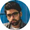 A man with a beard and glasses, wearing a dark blazer over a shirt, standing against a blue textured wall. he looks serious and is slightly facing to his right.