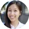 A close-up portrait of a smiling asian woman with long hair, wearing a white blouse, outdoors with soft-focused greenery in the background.