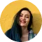 A woman with teal hair smiling, standing against a yellow background. she is wearing a denim jacket and has a vibrant lipstick shade.