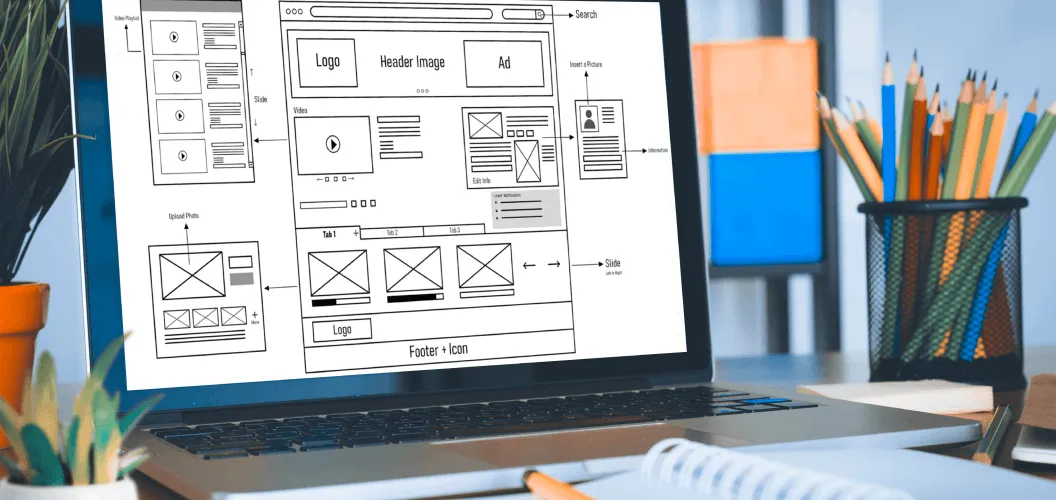 A laptop screen displaying a web design wireframe, positioned on a desk surrounded by office supplies such as pencils in a holder and a notebook.